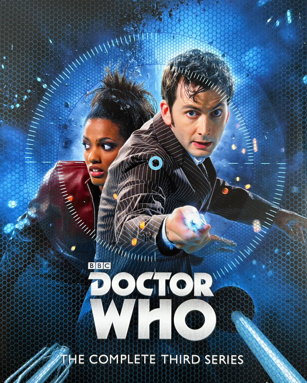 Front side of the flyer included with the Doctor Who Series 3 Blu-ray Steelbook, featuring a photo of David Tennant's Doctor wearing a pinstripe suit and holding his sonic screwdriver out in front of him, pointing towards us. Martha Jones, in her red leather jacket, presses herself against him from behind, as she looks fearfully to the side. The pair are viewed from the perspective of a Dalek, its gun and plunger just visible at the bottom of the image.