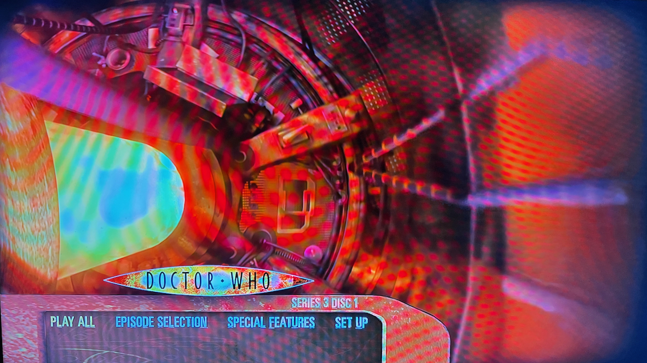 Blu-ray menu for Doctor Who Series 3, showing circular Tardis console viewed from above, bathed in a red light, while the central column of the console on the left is filled with a bright yellow light. The 4 options - Play All, Episode Selection, Special Features and Set Up - are displayed at the bottom of the screen, just below the Doctor Who logo.