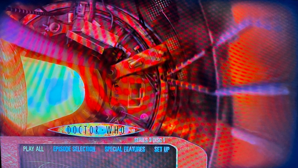 Blu-ray menu for Doctor Who Series 3, showing circular Tardis console viewed from above, bathed in a red light, while the central column of the console on the left is filled with a bright yellow light. The 4 options - Play All, Episode Selection, Special Features and Set Up - are displayed at the bottom of the screen, just below the Doctor Who logo.
