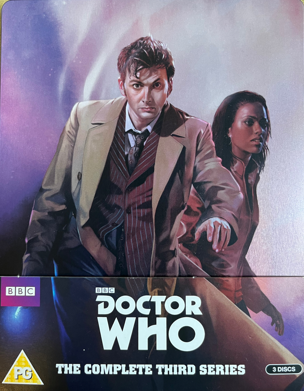 The front cover of the Doctor Who Series 3 Blu-ray steelbook, which is sitting in a cardboard pocket that has the Doctor Who logo on it. The steelbook cover shows David Tennant as the Doctor, wearing a light brown jacket over a pinstripe suit as he strides forward with his sonic screwdriver in his right hand and a serious, determined expression on his face. Martha Jones is behind him wearing a red leather jacket and glancing fearfully to the side.