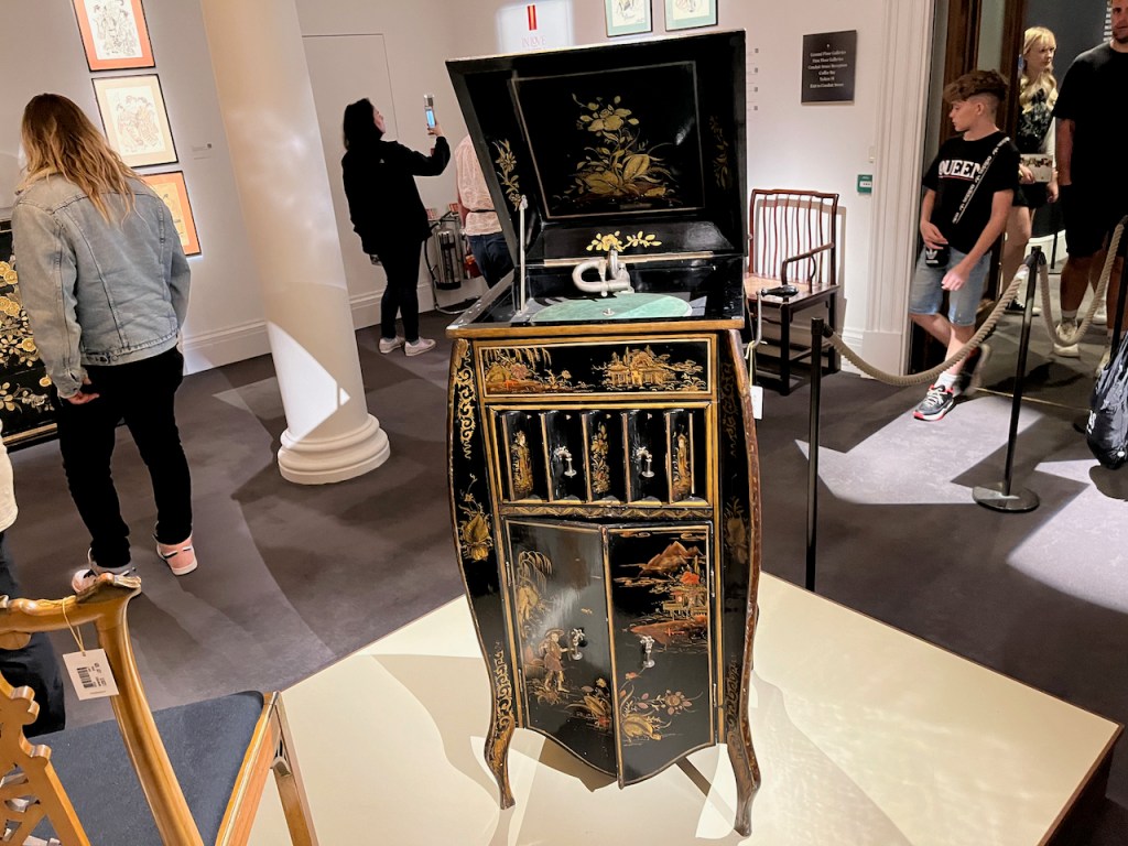 A tall, free-standing, ornately gold decorated, gramophone player, with the lid open to reveal the stylus inside.