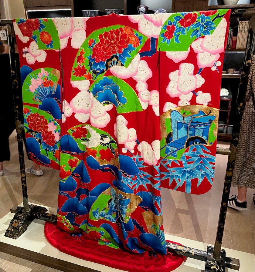The front of a brightly coloured Japanese kimono. The thick long-sleeved outer robe is decorated in stencil dye with branches of blossoming plum, stalks of bamboo and pine, interspersed with scattered fan-shaped images depicting ox-carts, peonies, pine, cranes in flight, chrysanthemums and irises.