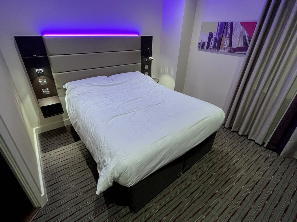 Double bed in the County Hall Premier Inn hotel, with a strip of purple light glowing from above the headboard on the wall.