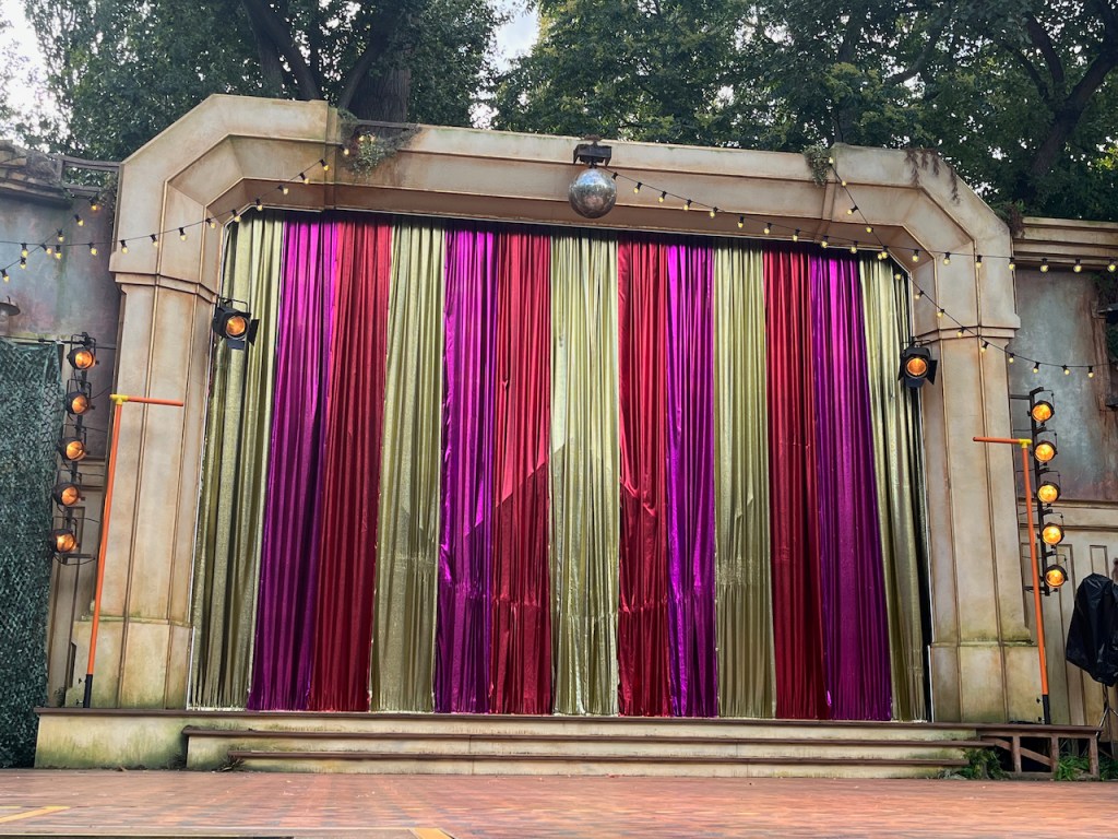 The curtain under a wide archway, at the back of the stage in the Regent's Park Open Air Theatre, for Sara Pascoe's show Success Story. The shiny material has vertical stripes of gold, pink and red repeating across it.