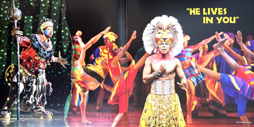 Double page spread from the programme for the Lion King musical, showing adult lion Simba, clutching a fist to his chest as he sings. The male actor's face is fully visible, with a lion mask attached to his forehead, with a thick mane around it. The top half of his chest his bare, below which his ornate golden outfit is decorated with African style markings. Behind him are lots of smiling dancers, kicking their legs high in the air, while large yellow letters at the top say He lives in you. An inset image to the left shows Rafiki the mandrill, wearing a very colourful outfit and face paint, and a crown on her head, looking around while holding a tall staff with a large sphere on the top of it.