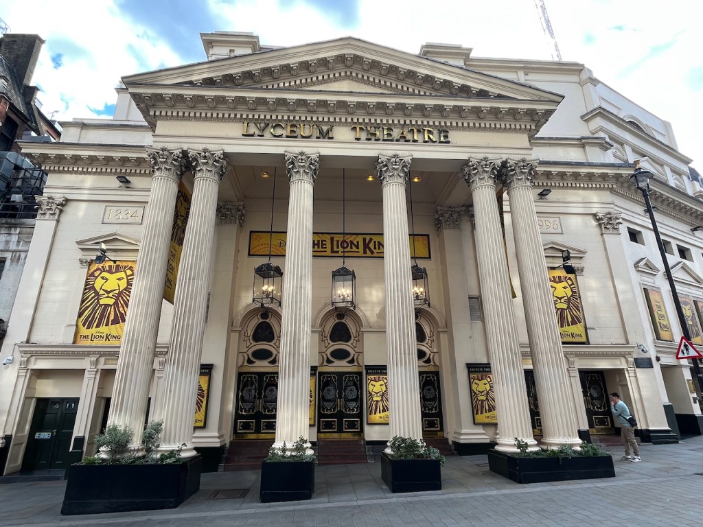 The Lyceum Theatre, with posters and signs for The Lion King. The entrance doors to the theatre are behind the 6 tall stone pillars across the front of the building.