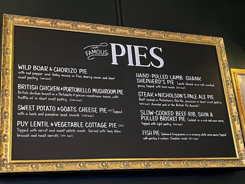 The Famous Pies menu on the wall in the Bear and Staff pub. The options include wild boar and chorizo pie, hand-pulled lamb shank shepherd's pie, British chicken and portobello mushroom pie, steak and Nicholson's pale ale pie, slow cooked beef rib, shin and pulled brisket pie, sweet potato and goats cheese pie, puy vegetable and lentil cottage pie, and fish pie containing salmon and king prawns.