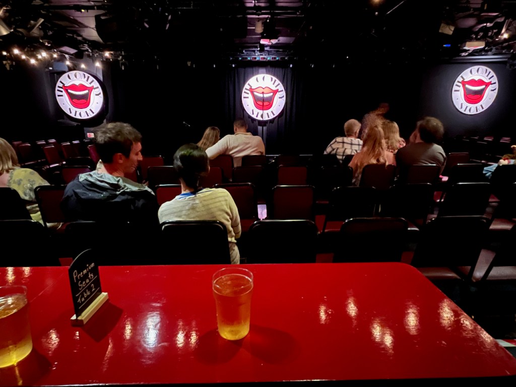 View towards the stage in the Comedy Store, from the red table at the front of the slightly raised Premium area, on which there are a couple of plastic glasses of cider, and a small black sign that has Premium Seats Table 2 written on it in white. There are 4 rows of standard seating ahead, between the table and the stage. The Comedy Store logo, with a big laughing mouth in the centre of a white circle with the venue name around its edge, is lit up on the left, centre and right of the stage.