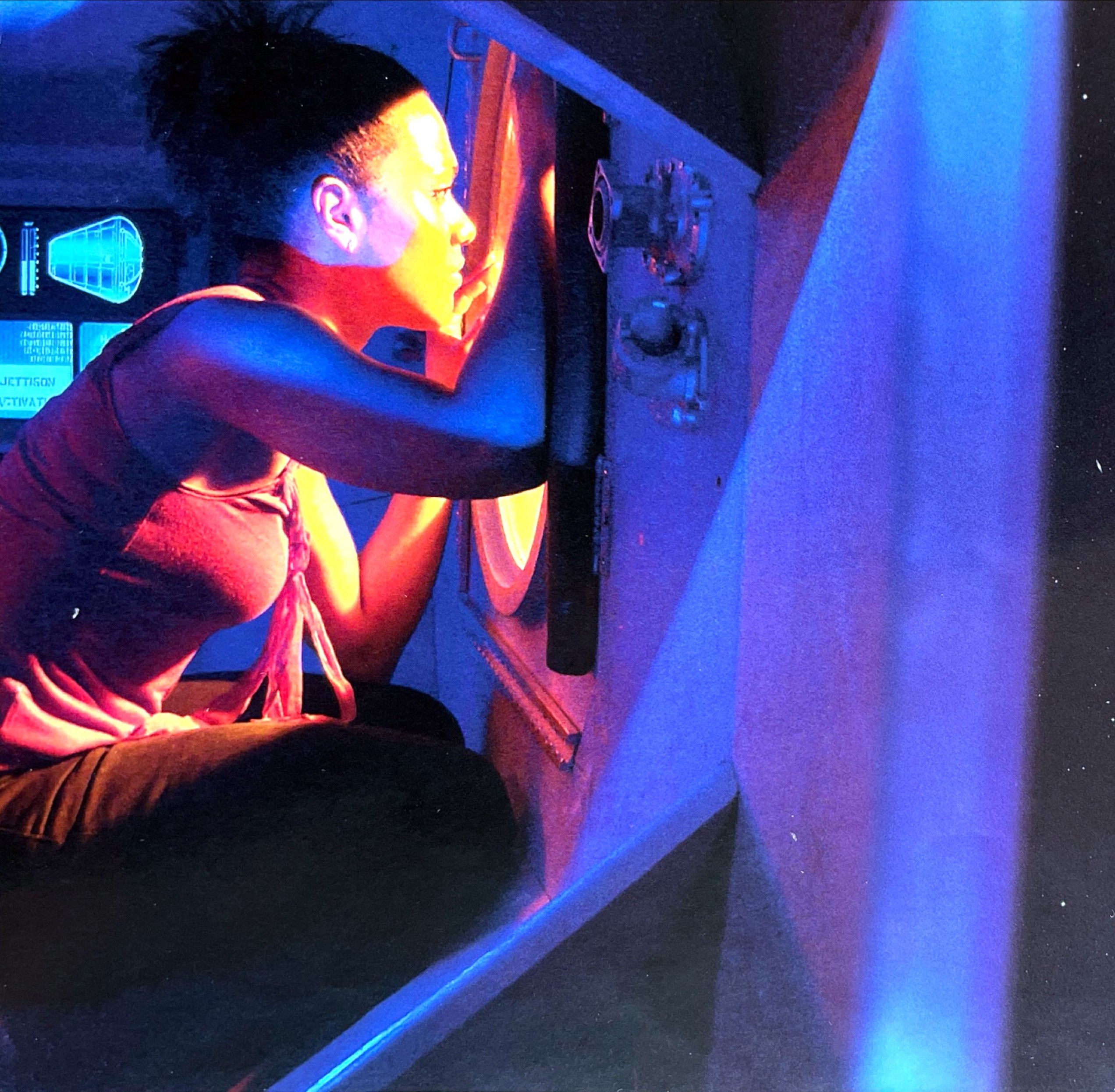 Martha Jones, her face illuminated with yellow light as she looks through the porthole window of a spaceship.