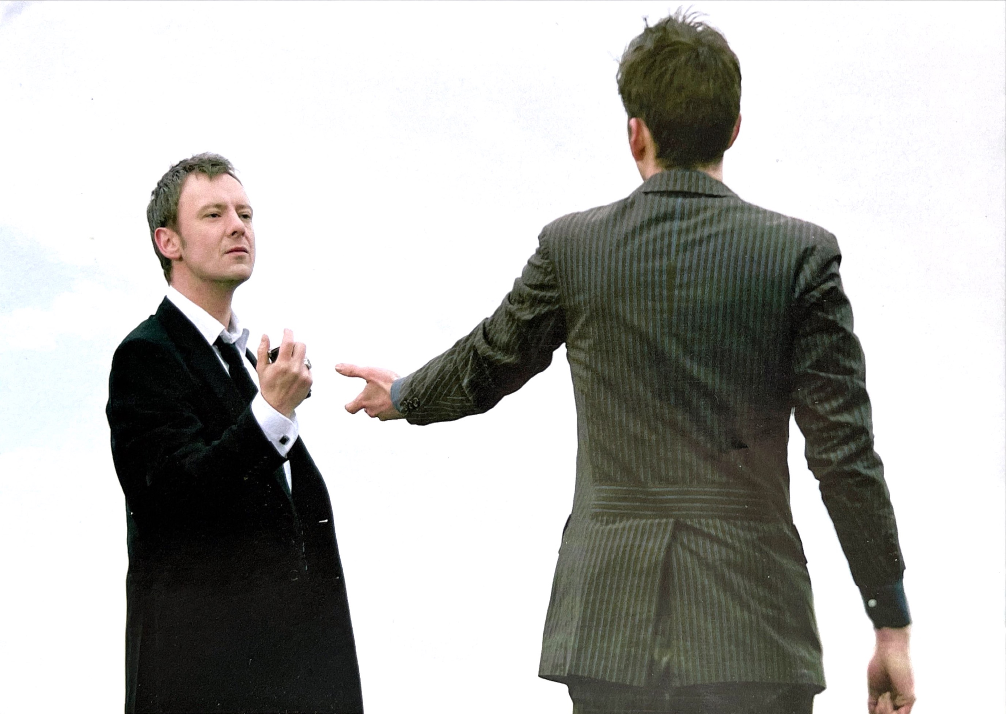 John Simm as the Master in a smart black suit and tie, and David Tennant as The Doctor in a pinstripe suit, standing and facing each other. The Doctor has his back to us, but we can see the defiant look on the Master's face as he clutches something in front of him in his right fist.