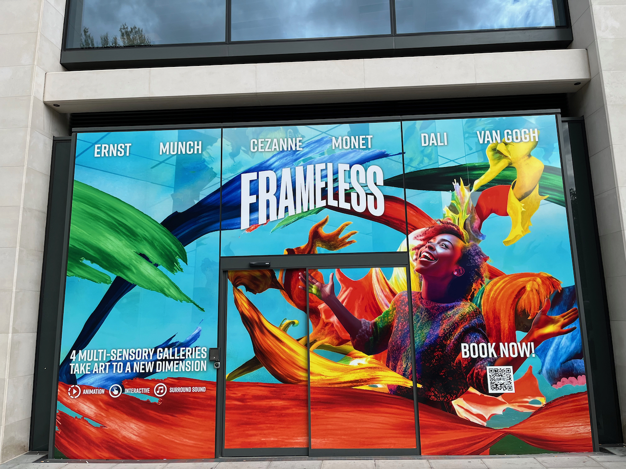 A large display on the outside of the Frameless gallery building, showing a woman gazing upwards with a smile, and her arms out to her sides, with large colourful paint strokes behind her. Across the top, white text gives the names of the artists Ernst, Munch, Cezanne, Monet, Dali and Van Gogh. At the bottom left of the display, white text says 4 multi-sensory galleries take art to a new dimension, with icons for animation, interactive and surround sound below it. And on the bottom right is a QR code with white text saying book now above it.
