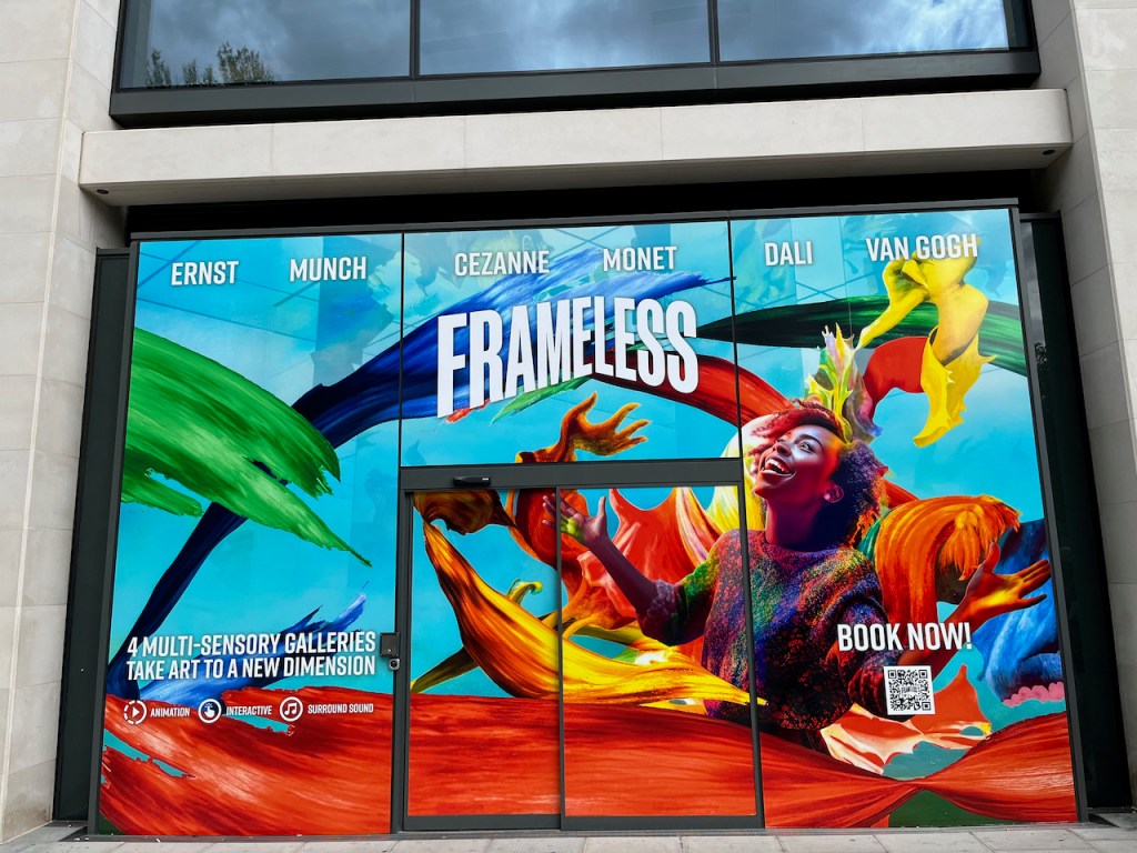 A large display on the outside of the Frameless gallery building, showing a woman gazing upwards with a smile, and her arms out to her sides, with large colourful paint strokes behind her. Across the top, white text gives the names of the artists Ernst, Munch, Cezanne, Monet, Dali and Van Gogh. At the bottom left of the display, white text says 4 multi-sensory galleries take art to a new dimension, with icons for animation, interactive and surround sound below it. And on the bottom right is a QR code with white text saying book now above it.