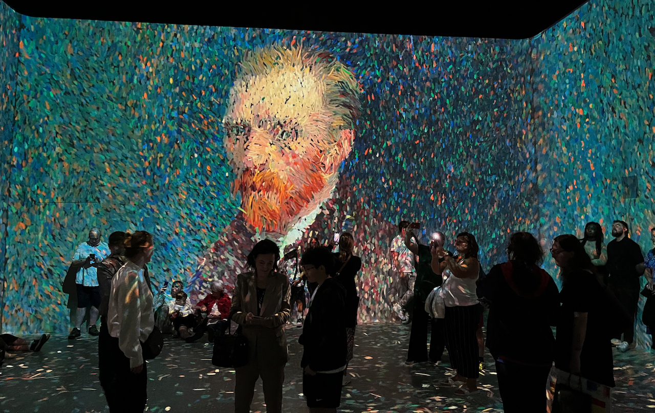 A self-portrait of Vincent Van Gogh, on a huge screen filling the wall of a room in the Frameless Gallery. The image is made up of lots of colourful, confetti-like, digital elements, some of which are still floating around on the floor, having been kicked towards the wall to make the image.
