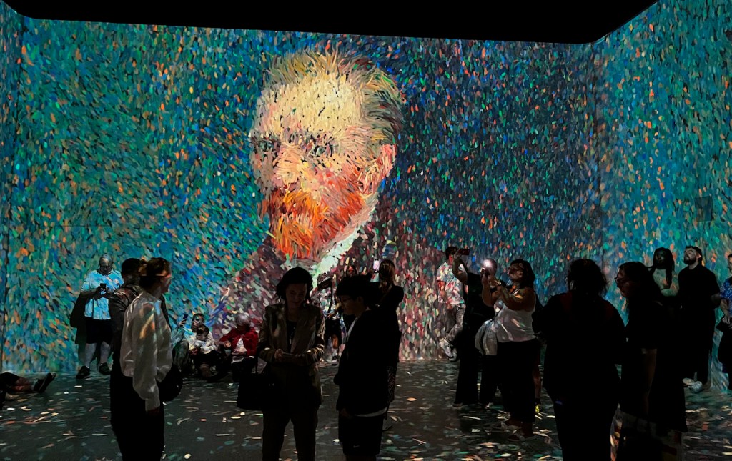 A self-portrait of Vincent Van Gogh, on a huge screen filling the wall of a room in the Frameless Gallery. The image is made up of lots of colourful, confetti-like, digital elements, some of which are still floating around on the floor, having been kicked towards the wall to make the image.