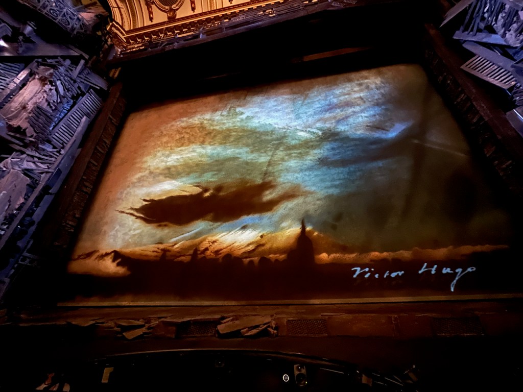 A large screen at the front of the stage before the start of Les Misérables, filled with a painting that's signed in white in the bottom right corner by Victor Hugo. As explained in the introductory notes from VocalEyes, it shows a glimpse of a small town at night, silhouettes of buildings and a Church spire. Overhead the sky is dark with flashes of white which stand out like the reflection of sun on water.