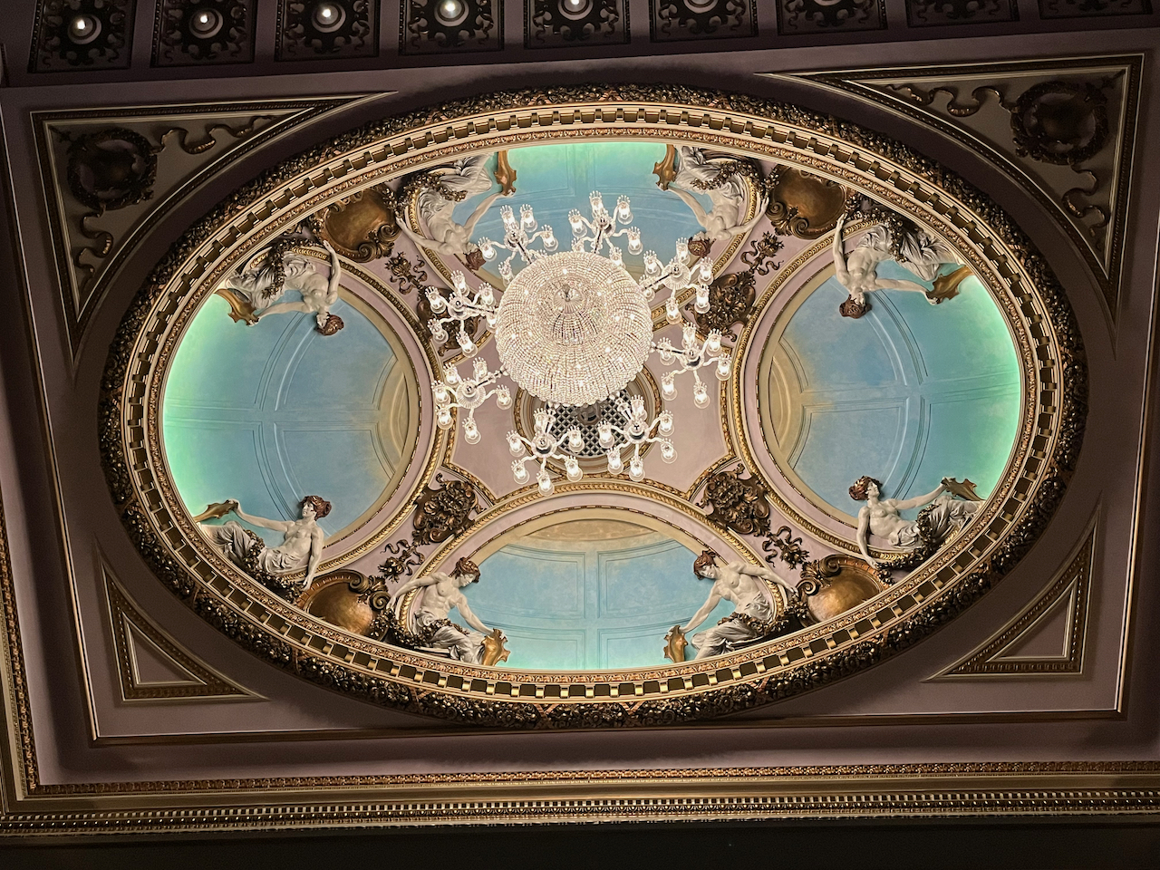 The ceiling of the Sondheim Theatre auditorium. Filling a rectangular framed space is a large oval, inside which, at 4 points around the inner edge, are topless female figures holding on to what appear to be leaves. These figures are in pairs, sitting back to back with each other. In the centre of space is an ornate chandelier, with a large glittering sphere in the centre. Branching off from that sphere are 8 mini chandeliers, each with 6 arms that each hold a single bulb.