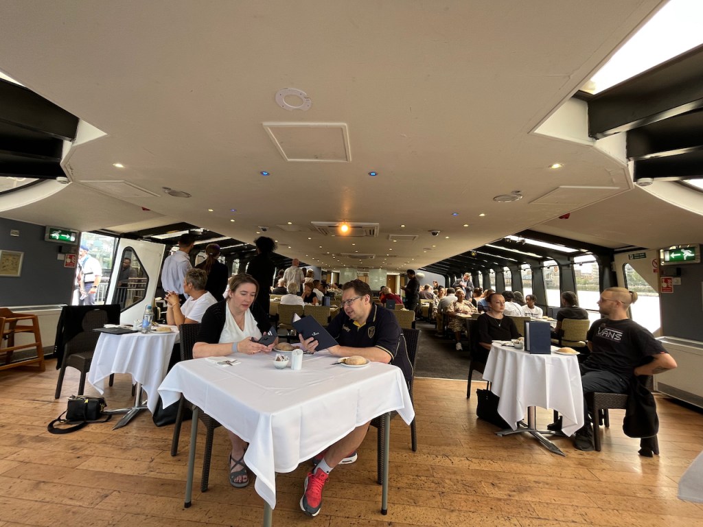 Dining room on the Millennium of London boat, viewed from our position at the front of the vessel. Some diners are sitting at 3 tables that are covered in white tablecloths on a wood-effect floor, while behind them in a carpeted area are many more diners seated at rows of plain tables.