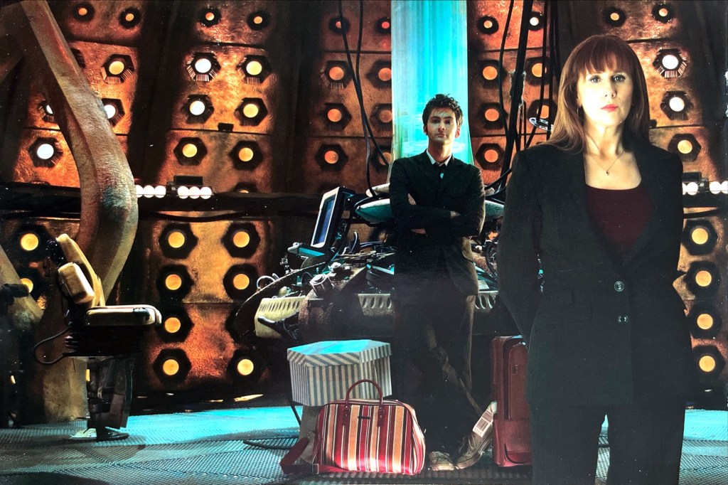 David Tennant's Doctor and Donna Noble standing inside the Tardis. Both are dressed in smart black outfits, the Doctor in a suit and Donna in a jacket over a top and trousers. The Doctor is leaning casually against the console with his arms folded and one ankle crossed over the other, and has Donna's suitcase and bags by his feet.