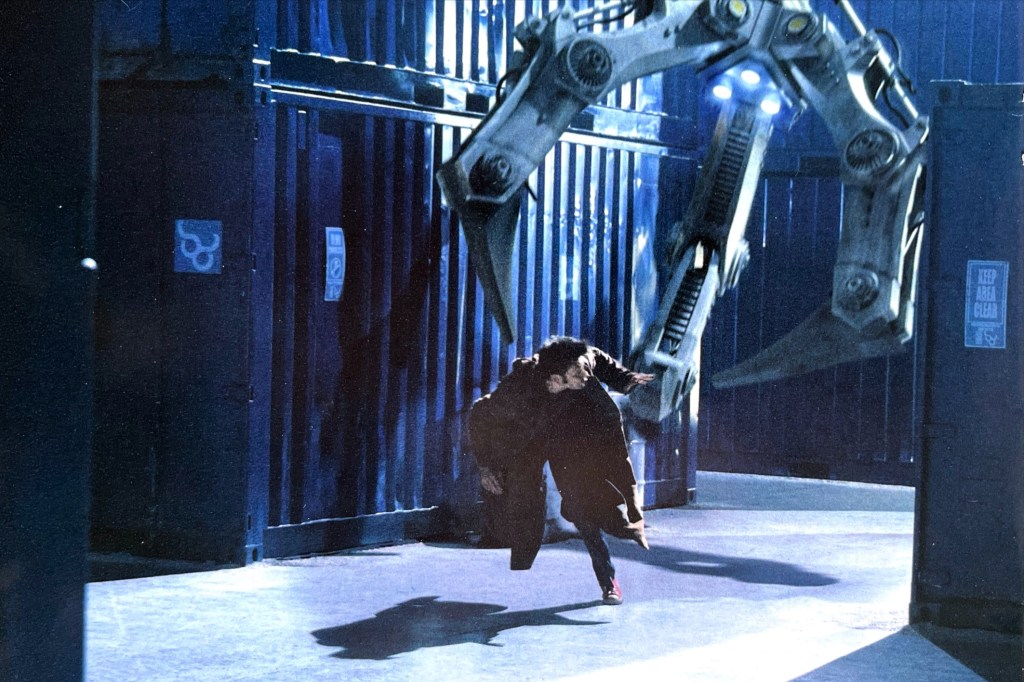 David Tennant as the 10th Doctor, running past large shipping containers and glancing behind him as a gigantic metal pincer overhead attempts to grab him.
