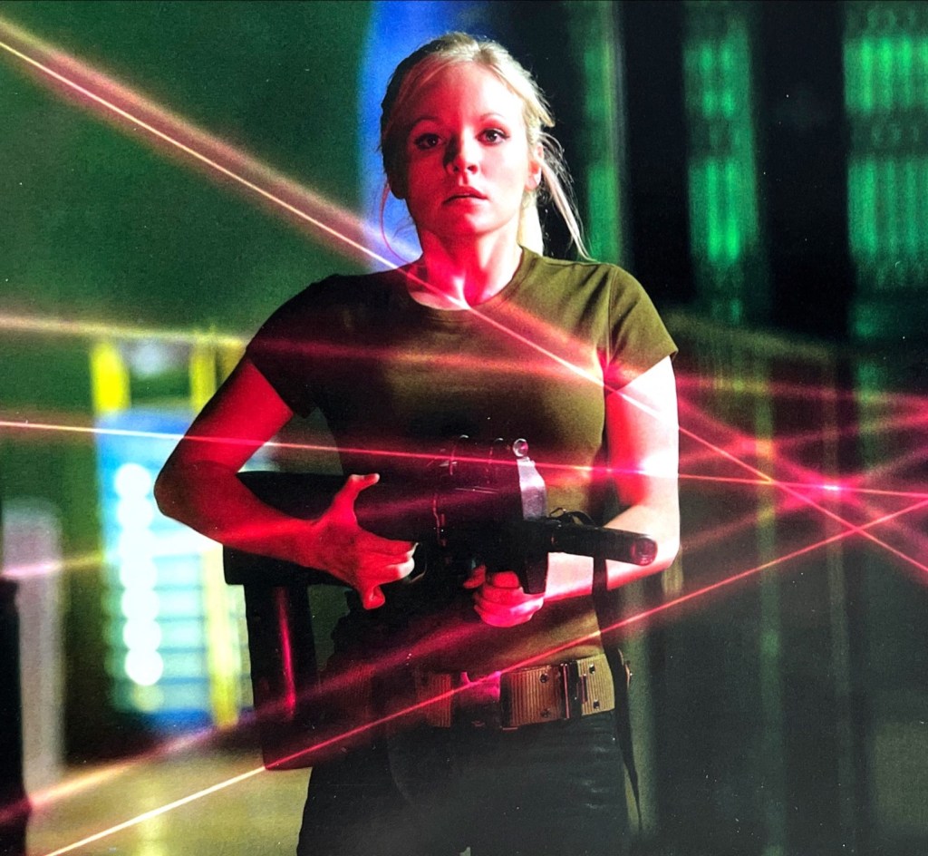 A pretty young blonde lady called Jenny, who was cloned from the DNA of the 10th Doctor. Wearing a dark green t-shirt and skintight black trousers, she holds a machine gun while looking in concern at the red lasers criss-crossing the pathway ahead of her.