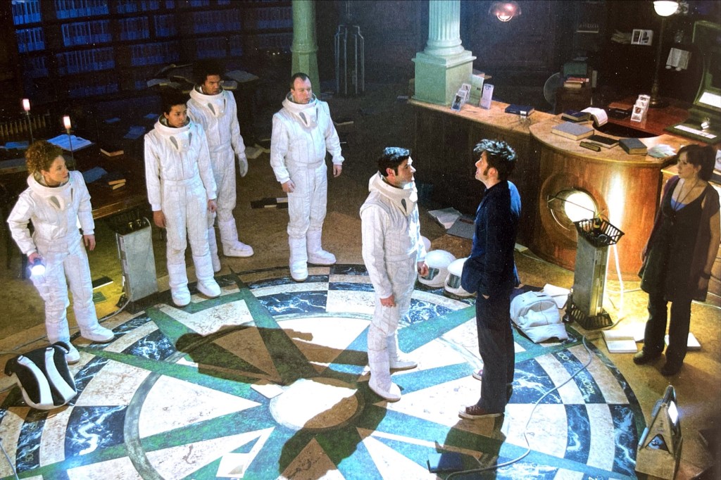 David Tennant's Doctor standing in a library with Donna Noble and a group of 5 people wearing white spacesuits. A man from the group stands in front of the Doctor. The Doctor is only casting one shadow on the ground on his left hand side. But the man has two shadows, one at the side and one behind him, despite being in the same lit area as the Doctor.