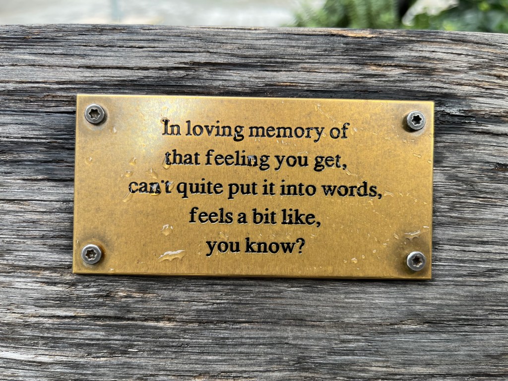 A gold bench plaque with black text that reads In loving memory of that feeling you get, can't quite put into words, feels a bit like, you know? The plaque is part of a series called In Loving Memory by Oliver Bragg.