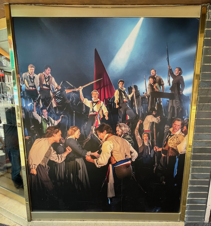 A poster in a gold frame on the outside of the Sondheim theatre, showing a photo of French revolutionaries facing us with defiance as they brandish their rifles, a couple of them holding them up in the air. At the bottom of the image, two men appear to be accosting a woman.