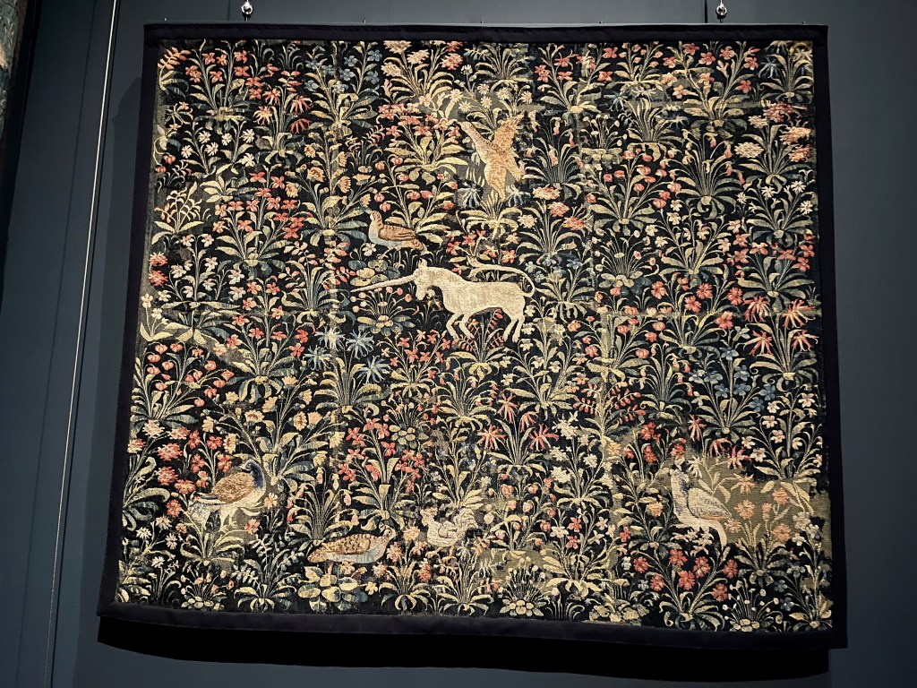 A large tapestry from around 1500 entitled Millefleurs with a Unicorn. Millefleurs means a thousand flowers, and there certainly many flowers, often on leafy stems, covering the tapestry. The small image of a unicorn in the centre represented chastity, since only a virgin might tame it, and invulnerability, as its horn was an antidote to poison.