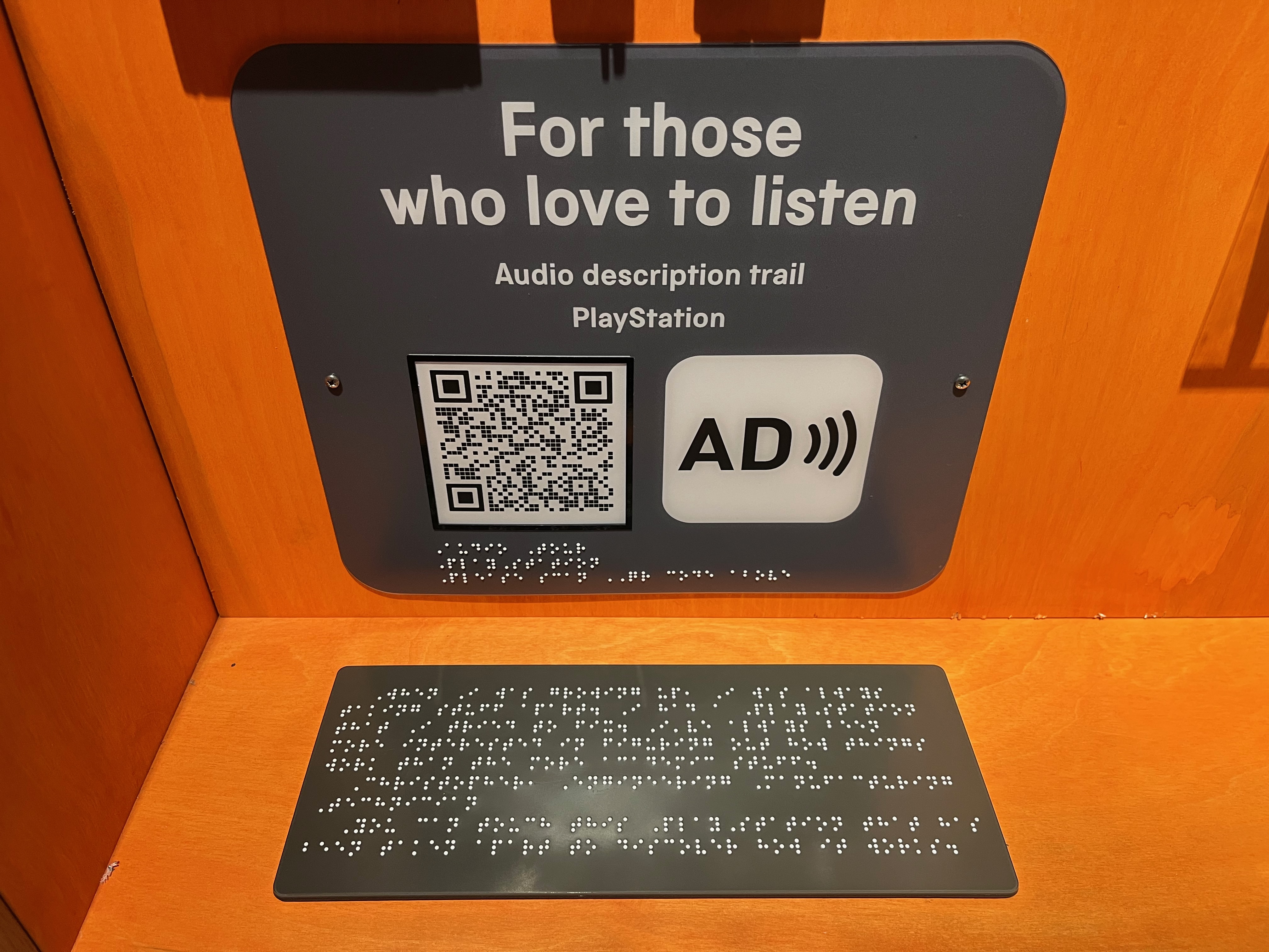 A dark grey panel with large white text that says For those who love to listen, with smaller text beneath that says Audio Description Trail, PlayStation. Below this is a QR code and the AD symbol, the latter consisting of the letters A and D together, with 3 curved lines radiating outwards to the right like sound waves. Below this is some braille text on a separate dark grey panel.