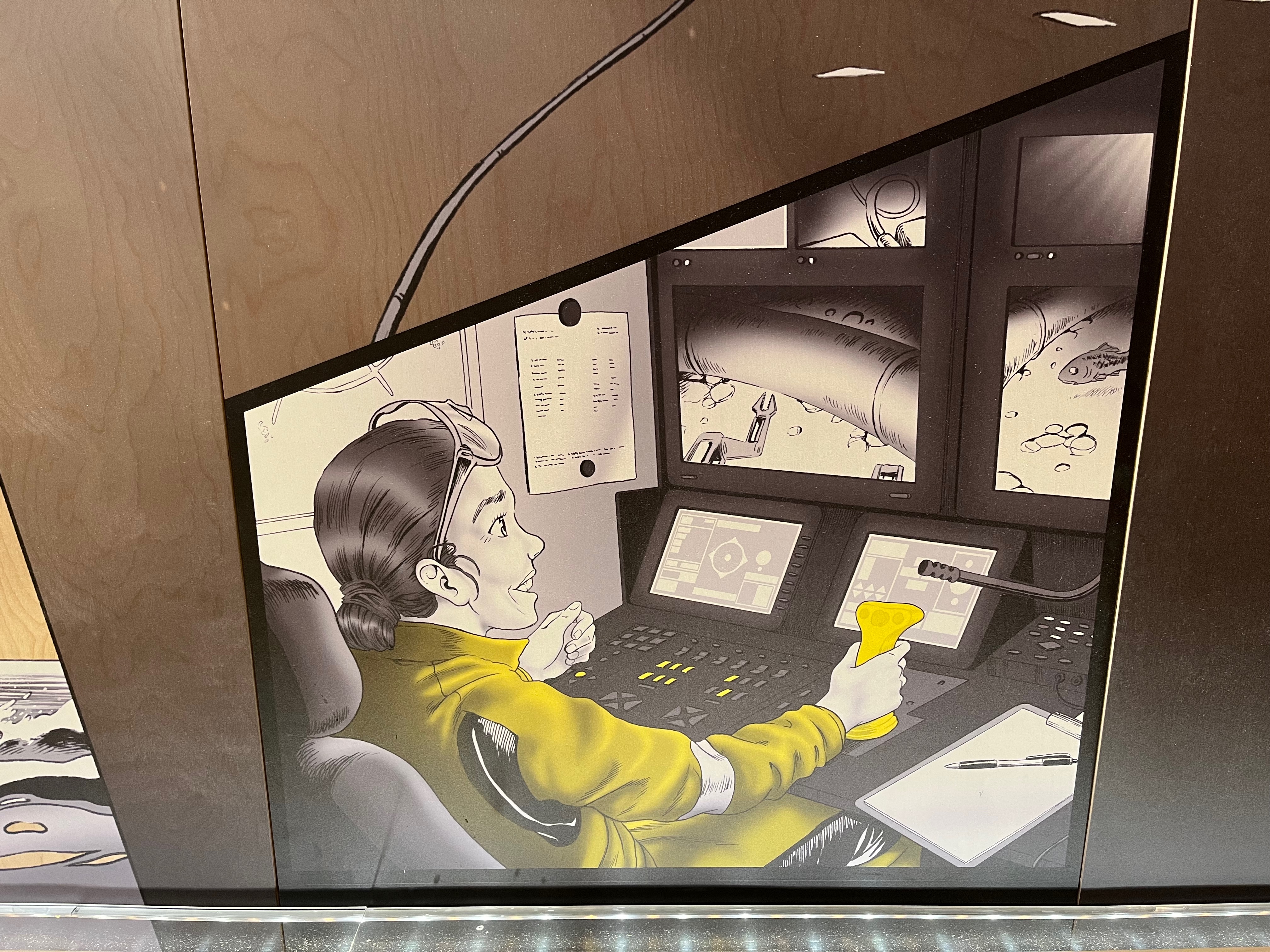 A large cartoon image of a smiling technician sitting in front of a console, using a joystick to control a robotic arm on a screen in order to fix an underwater pipe, as a fish looks on in confusion. The imagine is predominantly black and white, but the technician's jacket, the joystick and a few of the lights on the console are yellow.