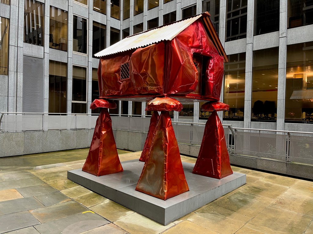 Sculpture called The Granary by Jesse Pollock. It is a life-sized sculpture of a traditional English grain store, consisting of a small hut with a corrugated iron roof and diagonal criss-cross bars on the small windows on the door and on the side. The hut is perched on 4 legs that have very large pyramid-shaped feet. The sculpture is finished in a pearlescent candy orange, and is designed to look like the hut has been a bit battered out of shape.