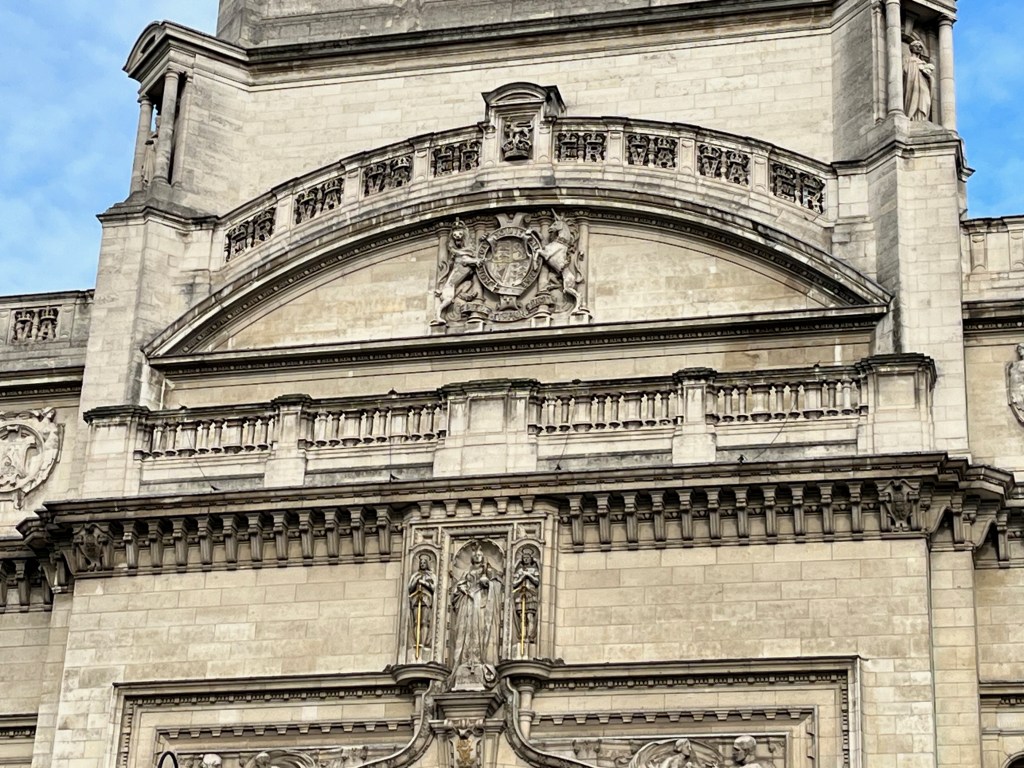 Close-up of sculpted figures high above the entrance on the front of the Victoria and Albert Museum. Two horses form part of a coat of arms, while further down 2 guards stand next to Queen Victoria.