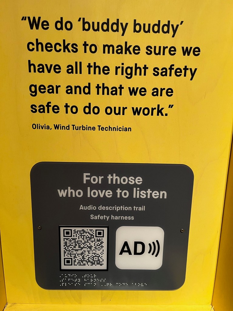 A yellow panel featuring a quote in large black text from Olivia, a wind turbine technician, who says We do buddy buddy checks to make sure we have all the right safety gear and that we are safe to do our work. Below that quote, a dark grey panel has large white text that says For those who love to listen, with smaller text beneath that says Audio Description Trail, Safety harness. Below that, still in the grey panel, is a QR code and the AD symbol, the latter consisting of the letters A and D together, with 3 curved lines radiating outwards to the right like sound waves.