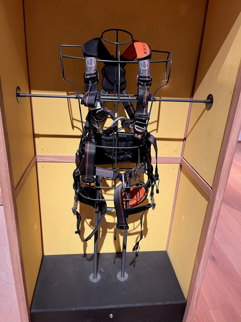 A large black safety harness used by wind turbine technicians, attached to metal bars in a yellow display case for people to touch.