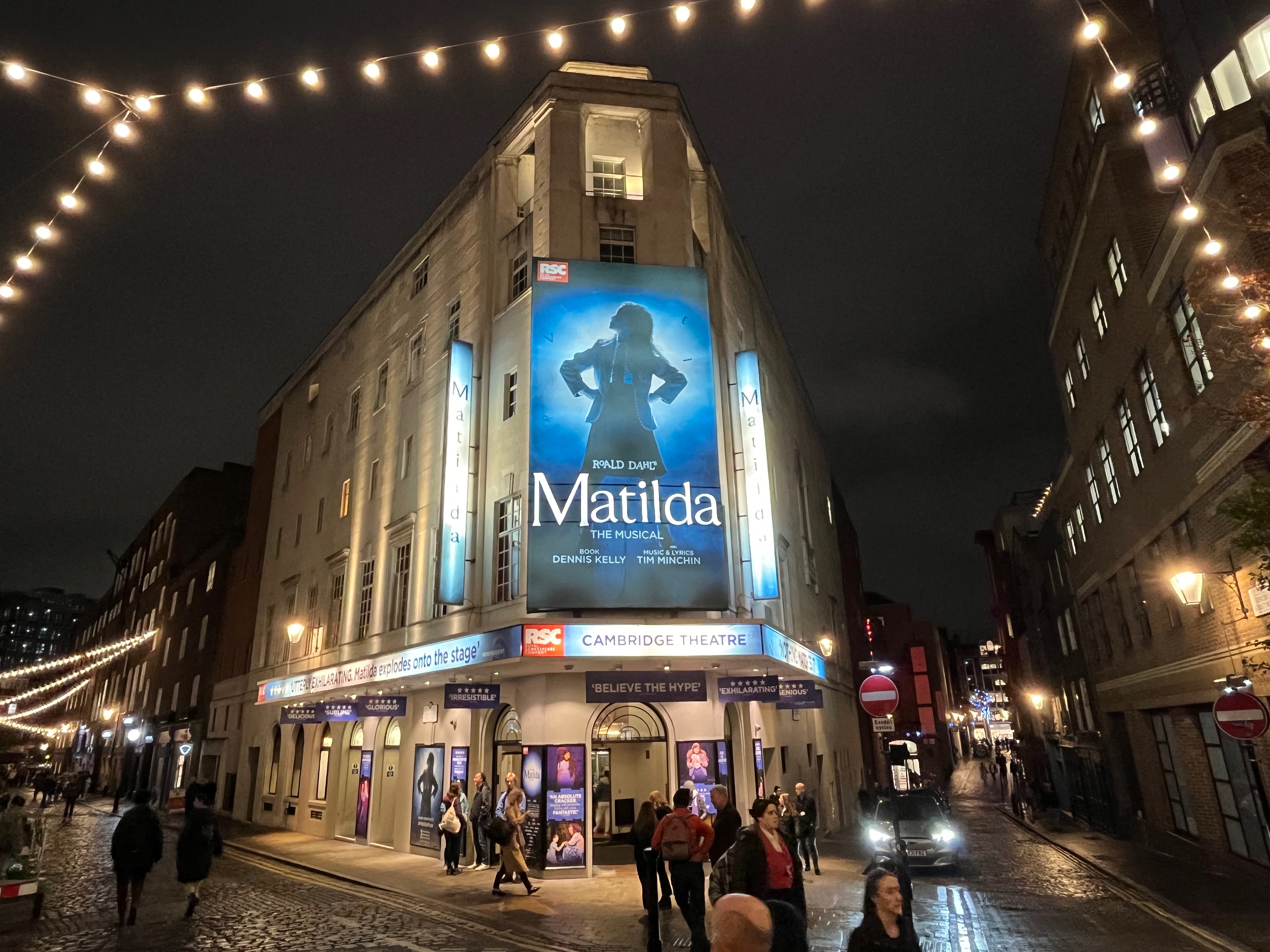 The Cambridge Theatre, with the large poster for Matilda The Musical dominating the building above the entrance on the corner, showing a silhouette of Matilda standing defiantly with her hands on her hips.