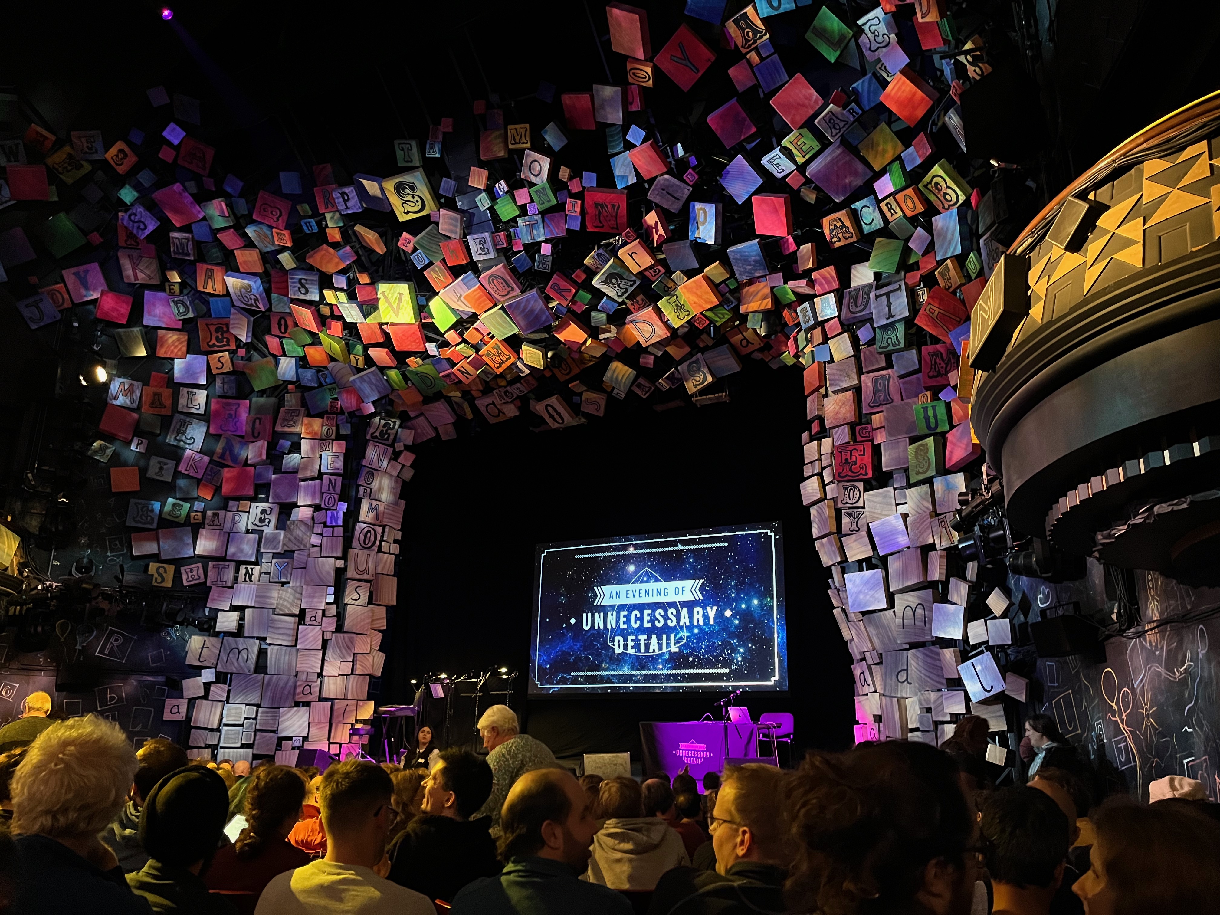 View towards the stage in the Cambridge Theatre for An Evening Of Unnecessary Detail, with the show's logo on the big screen at the back of the stage. The stage is surrounded by the many colourful alphabet blocks that normally frame the set for Matilda The Musical.