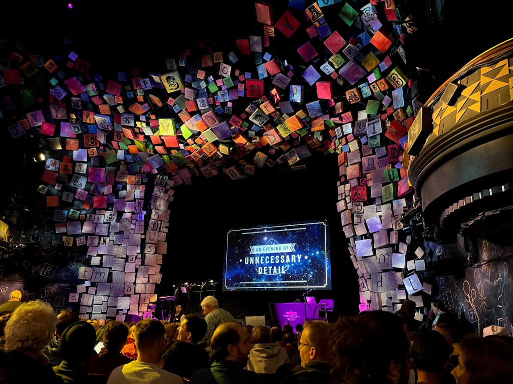 View towards the stage in the Cambridge Theatre for An Evening Of Unnecessary Detail, with the show's logo on the big screen at the back of the stage. The stage is surrounded by the many colourful alphabet blocks that normally frame the set for Matilda The Musical.