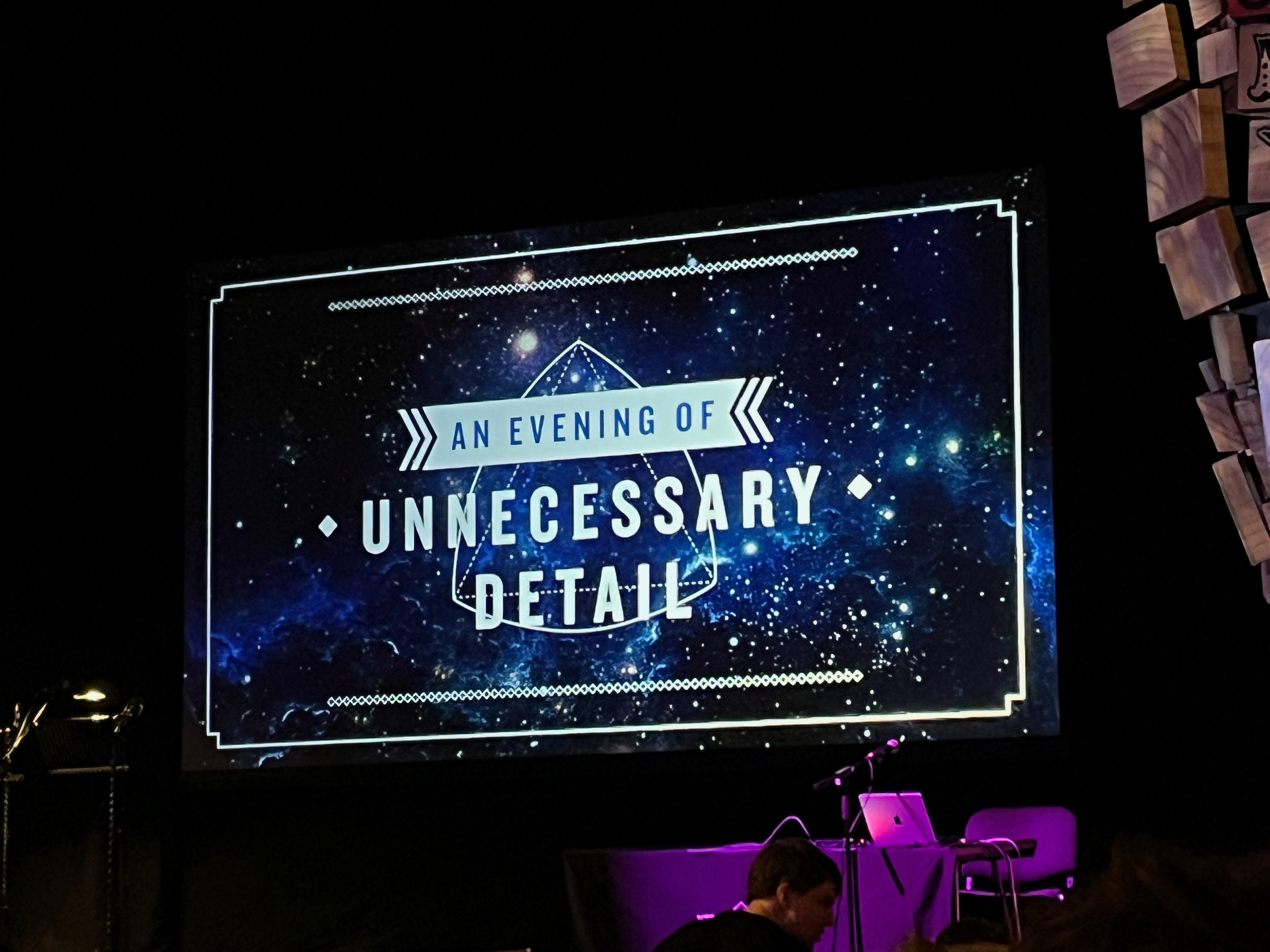 Close-up of the screen on stage in the Cambridge Theatre, showing the logo for An Evening Of Unnecessary Detail. The words An Evening Of are in dark lettering on a white bar, while the words Unnecessary Detail are in much larger white text beneath it, against a star-filled backdrop of space.