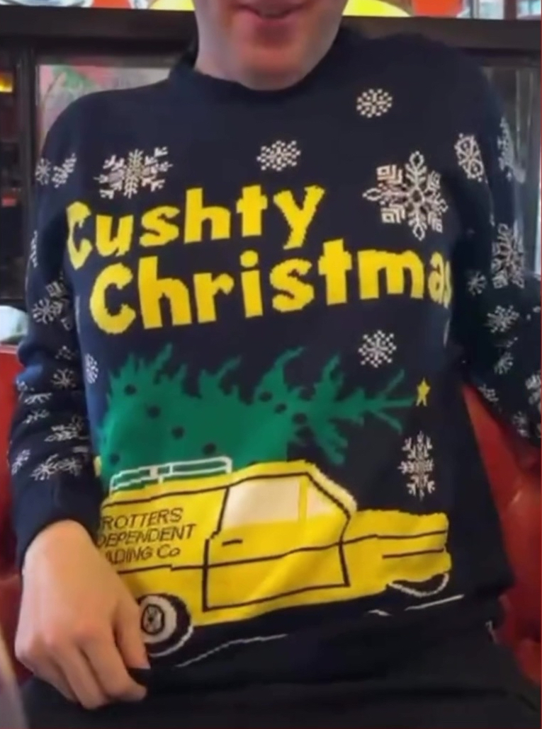 Glen in Bill's restaurant wearing an Only Fools And Horses Christmas jumper. The navy jumper has the words Cushty Christmas in large yellow letters above an image of a yellow three-wheeled Robin Reliant van, with Trotters Independent Trading Co written on the side and a green Christmas tree laid on its side on the roof. White snowflakes in different sizes decorate the rest of the jumper.