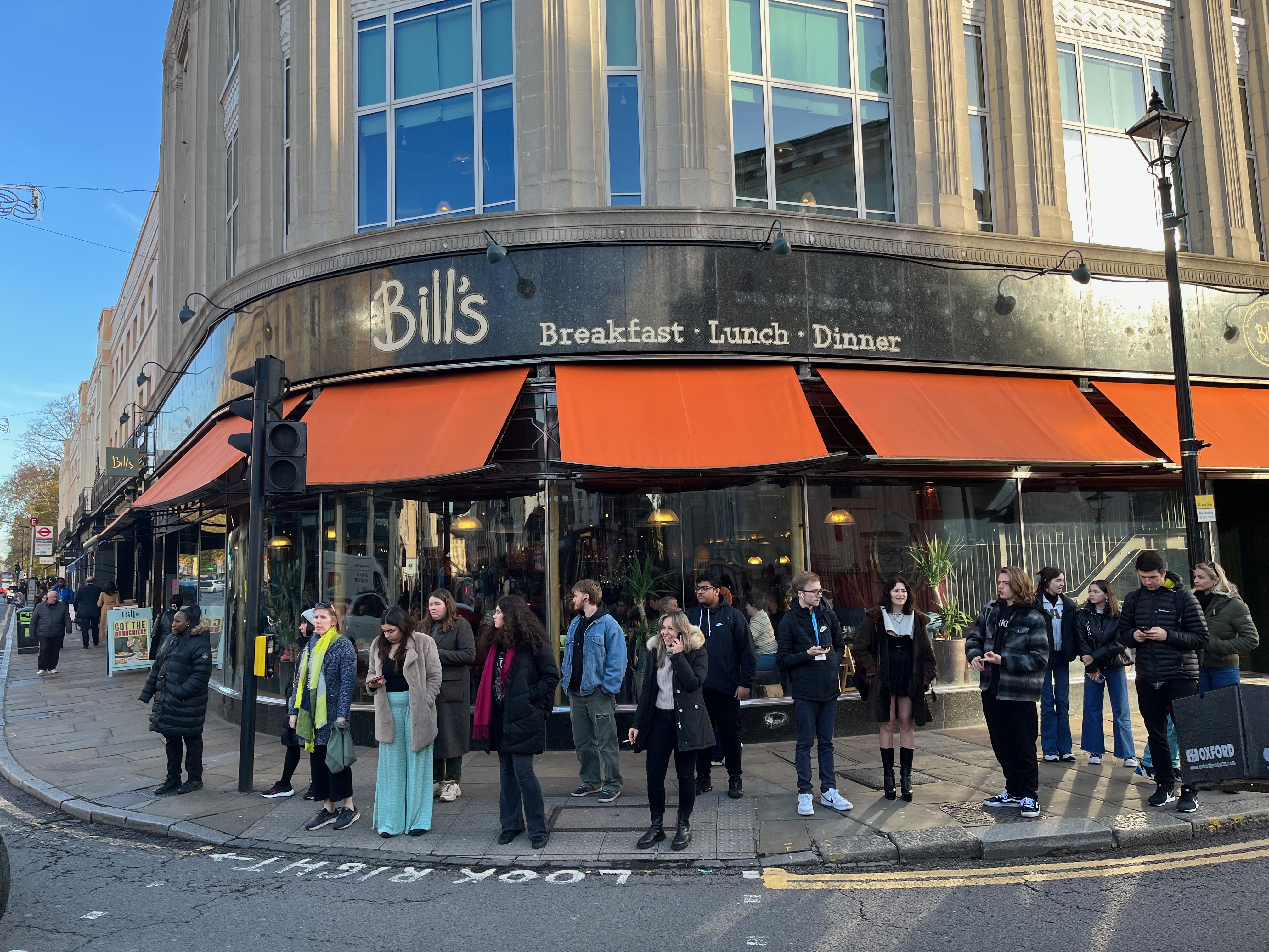 The exterior of Bill's restaurant on the corner of a junction in Greenwich. Small orange canopies hang over the windows on the ground floor. Above the canopies, in white lettering on a black background, is the logo for Bill's alongside the words Breakfast, Lunch, Dinner.