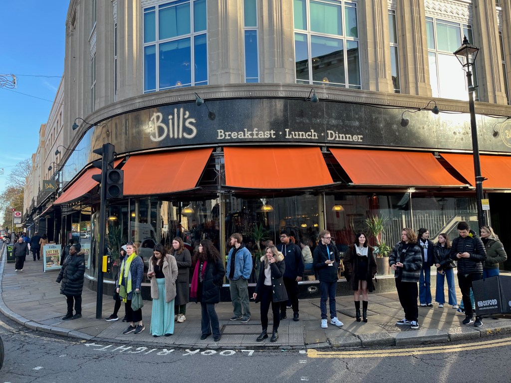 The exterior of Bill's restaurant on the corner of a junction in Greenwich. Small orange canopies hang over the windows on the ground floor. Above the canopies, in white lettering on a black background, is the logo for Bill's alongside the words Breakfast, Lunch, Dinner.