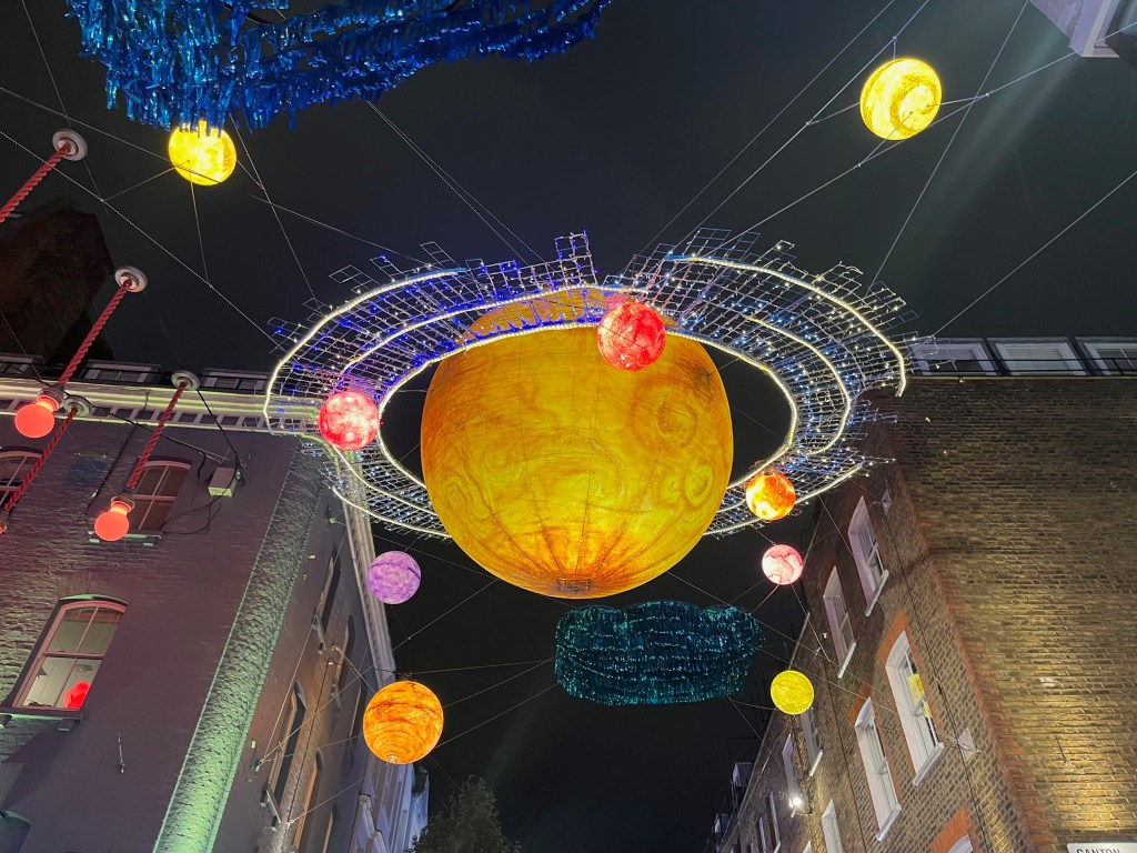 A large orange planet, with a glittering ring of lights around it, hanging above the crossroads in Carnaby Street. Lots of smaller planets or moons, in various bright colours, are hovering around it.