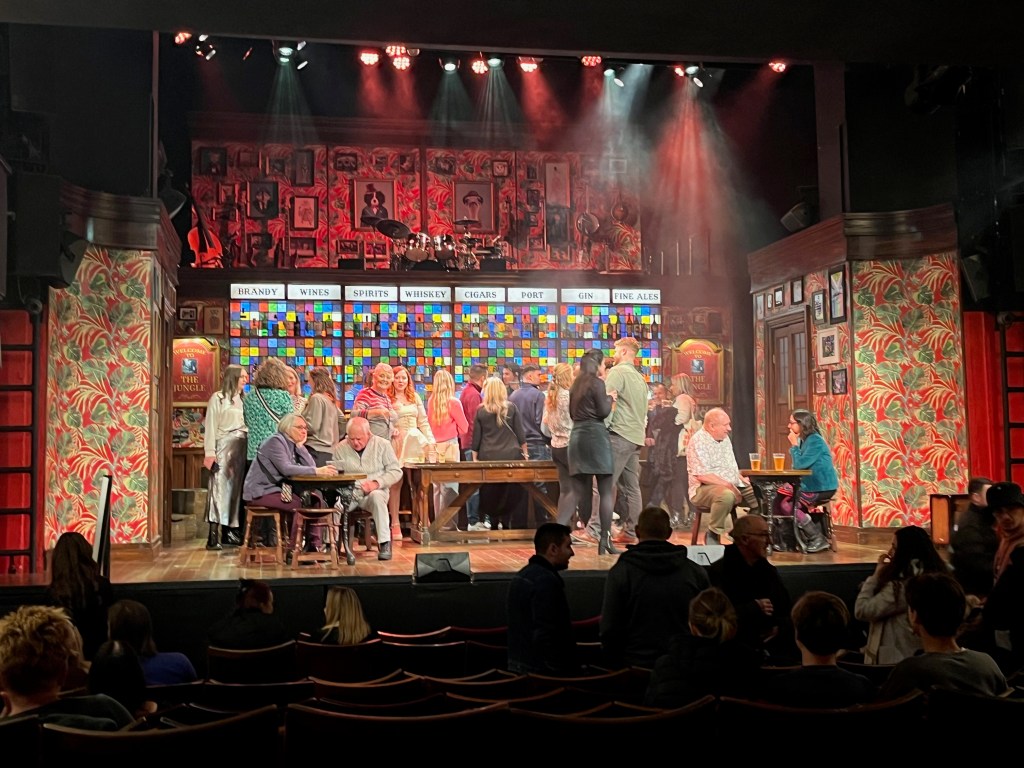 The stage for The Choir Of Man. It's set out like a pub, with a bar across the back of the space. Audience and cast members are on stage ordering from the bar or sitting at small tables before the show starts. Signs either side of the bar say Welcome to The Jungle. The backdrop of the bar is very colourful, and running across the top are white signs with black text saying Brandy, Wines, Spirits, Whiskey, Cigars, Port, Gin and Fine Ales. On an upper floor immediately above the bar is where the 4-piece backing band perform, so the drum kit is visible.