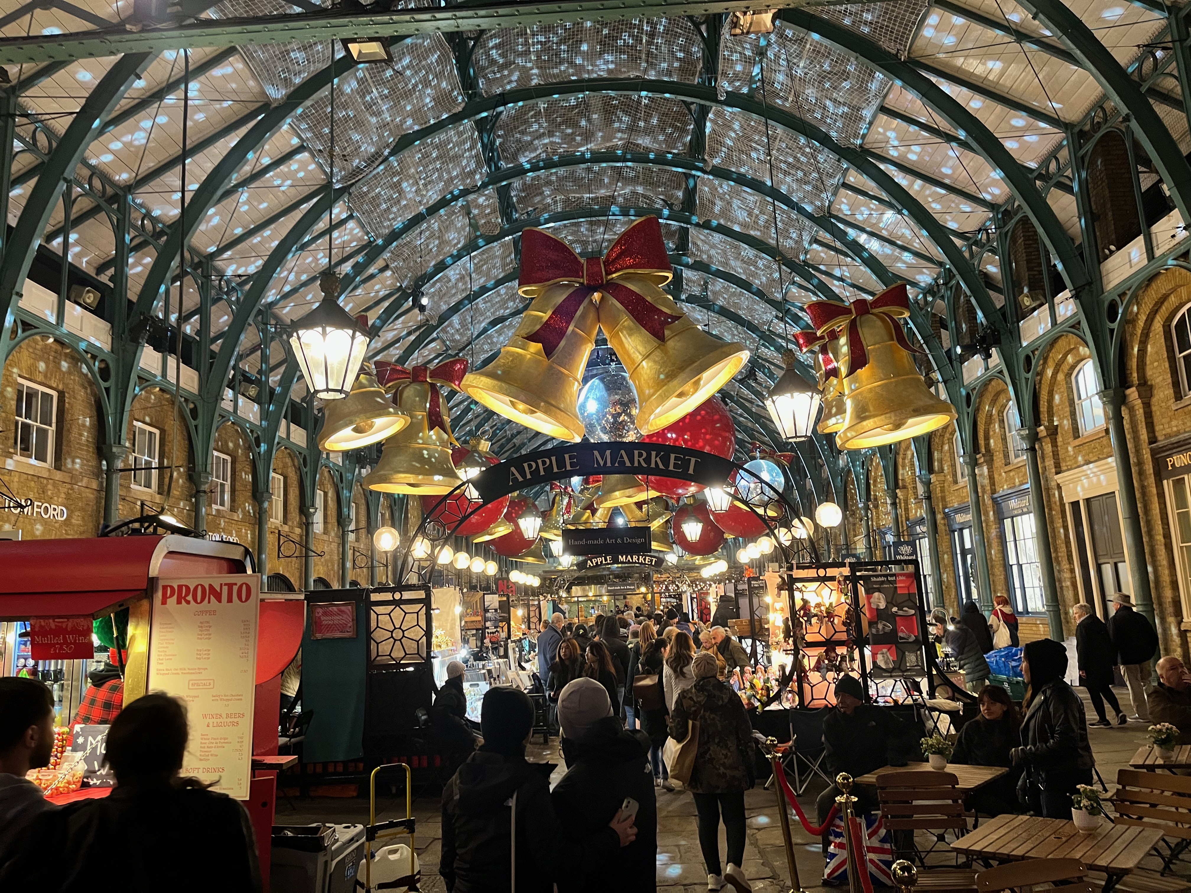 Large golden bells red glittery bows on top hang from the arched ceiling inside Covent Garden Market, above the throng of people browsing the stalls. Hanging mirror balls scatter moving speckles of light all over the ceiling as well, and a few lamps from the ceiling throughout the market.