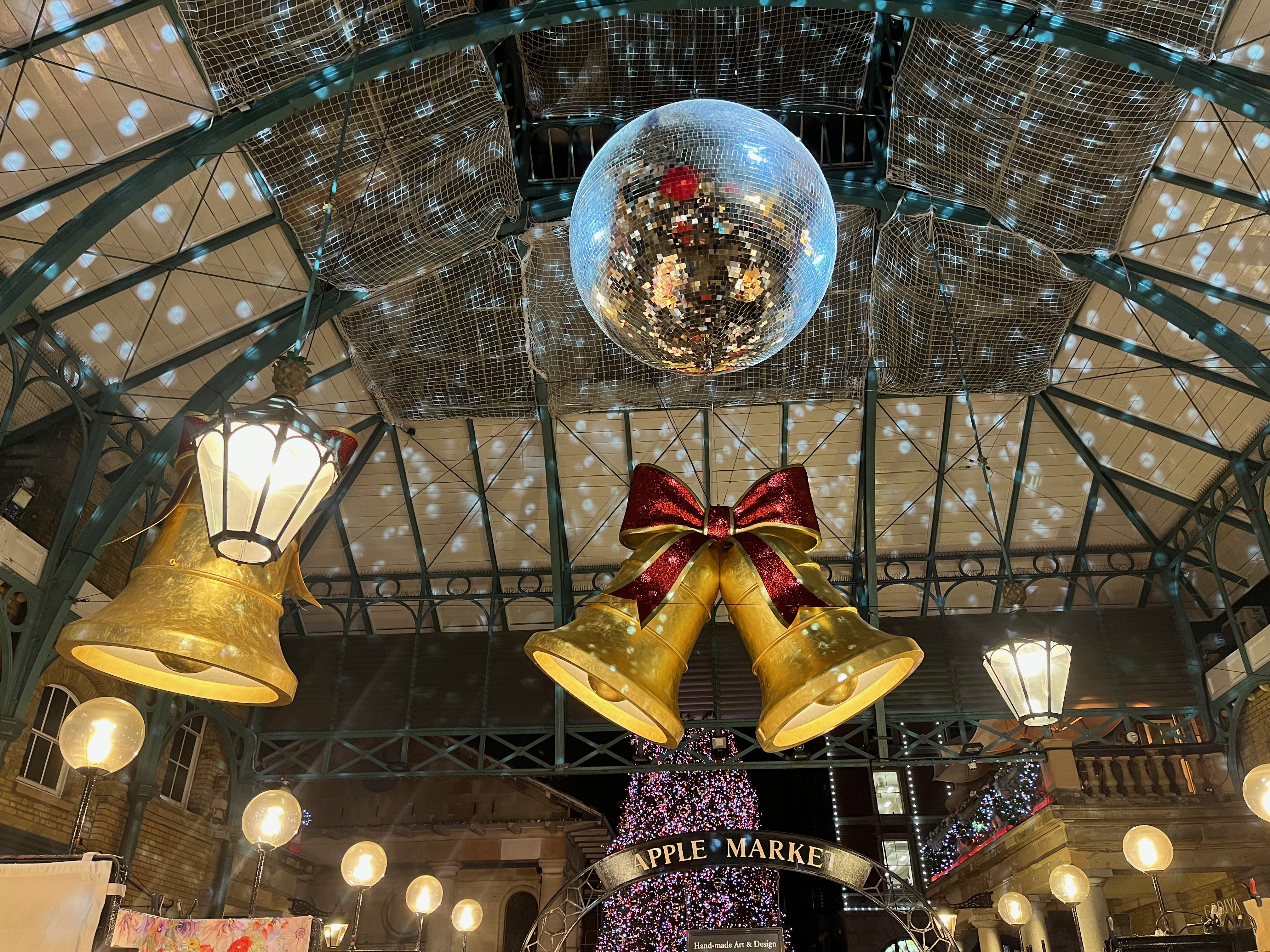 A mirror ball projecting spots of light on to the ceiling of Covent Garden Market. Behind it, just above the entrance, are two bells with a glittering red bow on top above a sign saying Apple Market. Behind that, outside the Market, part of a large Christmas tree is visible.