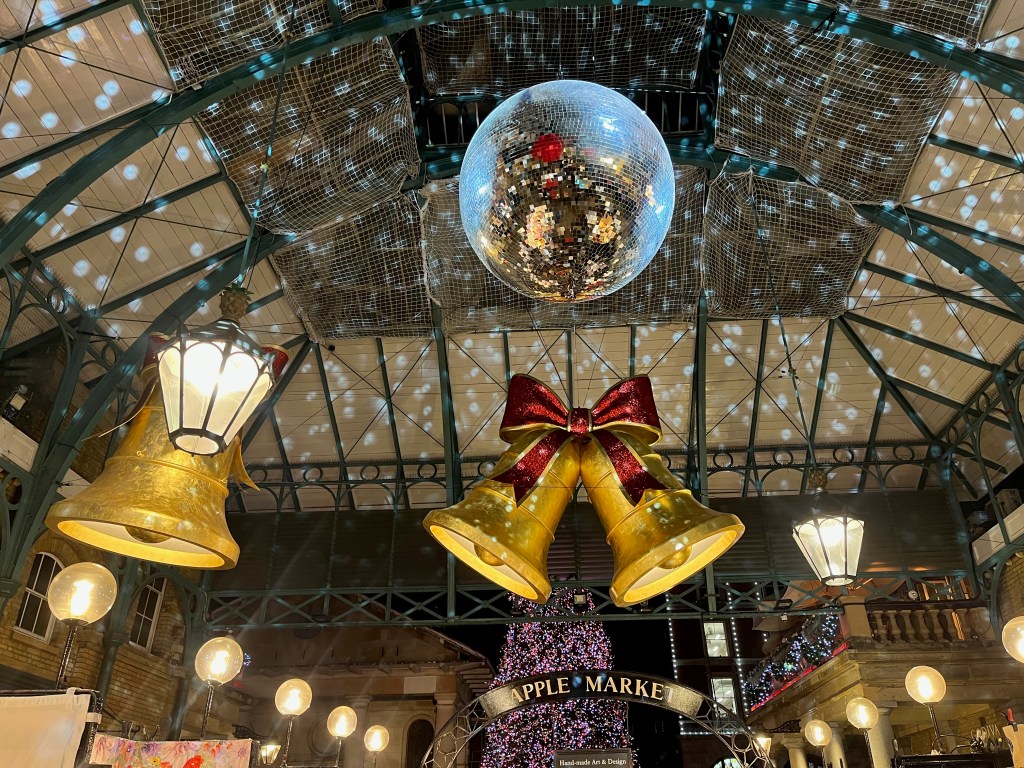 A mirror ball projecting spots of light on to the ceiling of Covent Garden Market. Behind it, just above the entrance, are two bells with a glittering red bow on top above a sign saying Apple Market. Behind that, outside the Market, part of a large Christmas tree is visible.