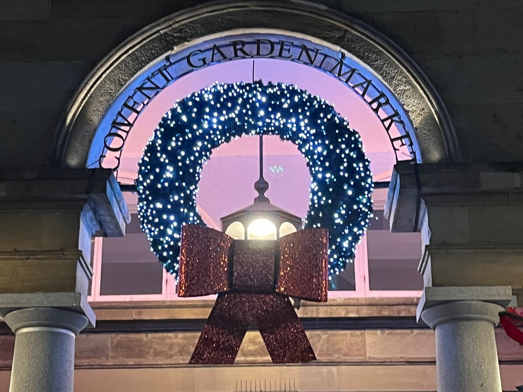 A wreath covered in lights, and with a red glittery bow underneath, attached to an arched window that has the words Covent Garden Market around the top of it.