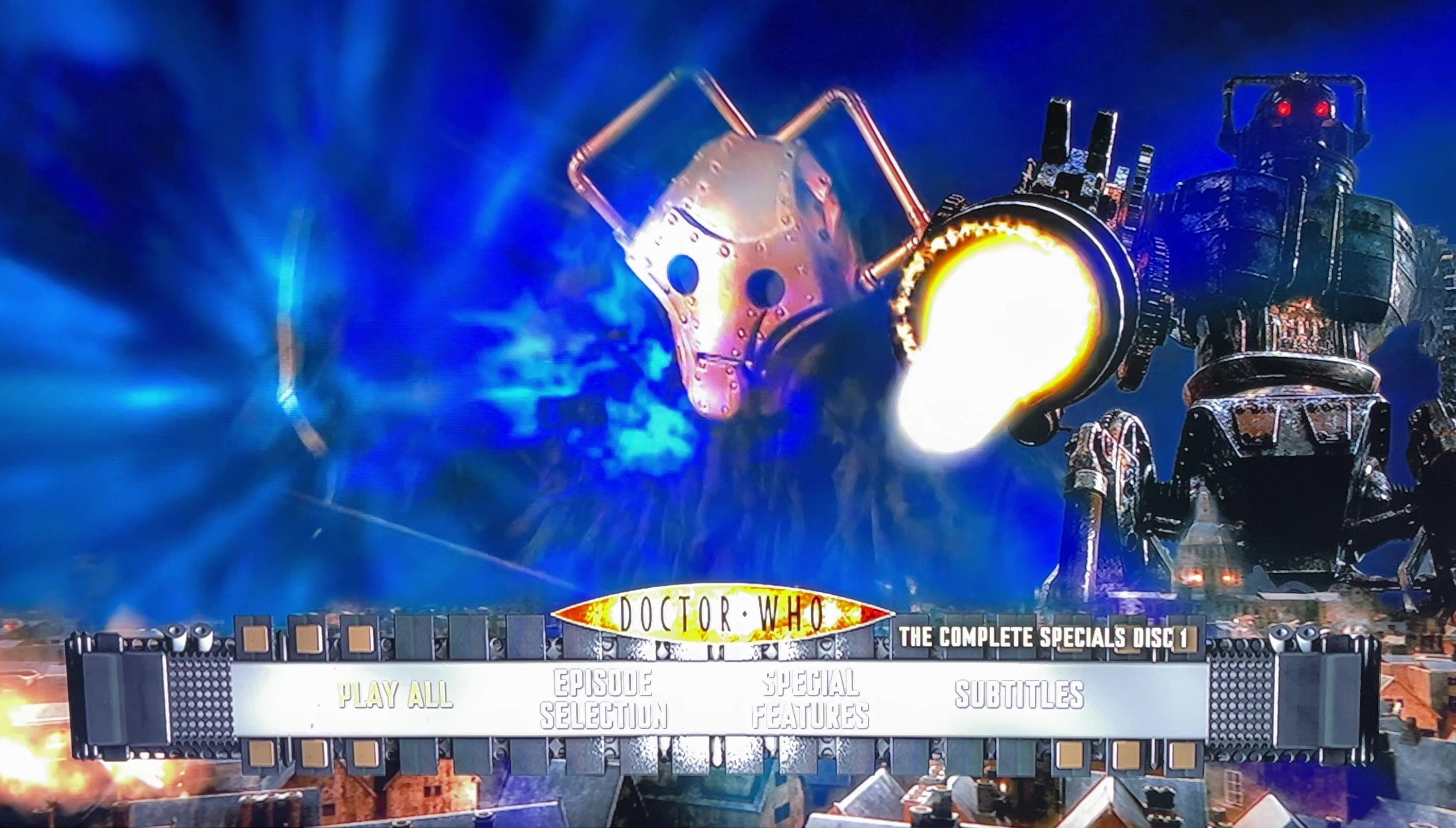 The menu for Disc 1 of the Doctor Who 2008 to 2010 specials Blu-ray steelbook. The screen shows a Cybershade, a small metal dog-like beast, next to the CyberKing, a huge robot, the latter firing a big fireball. The menu options are across the bottom of the screen, with the Doctor Who logo just above them.