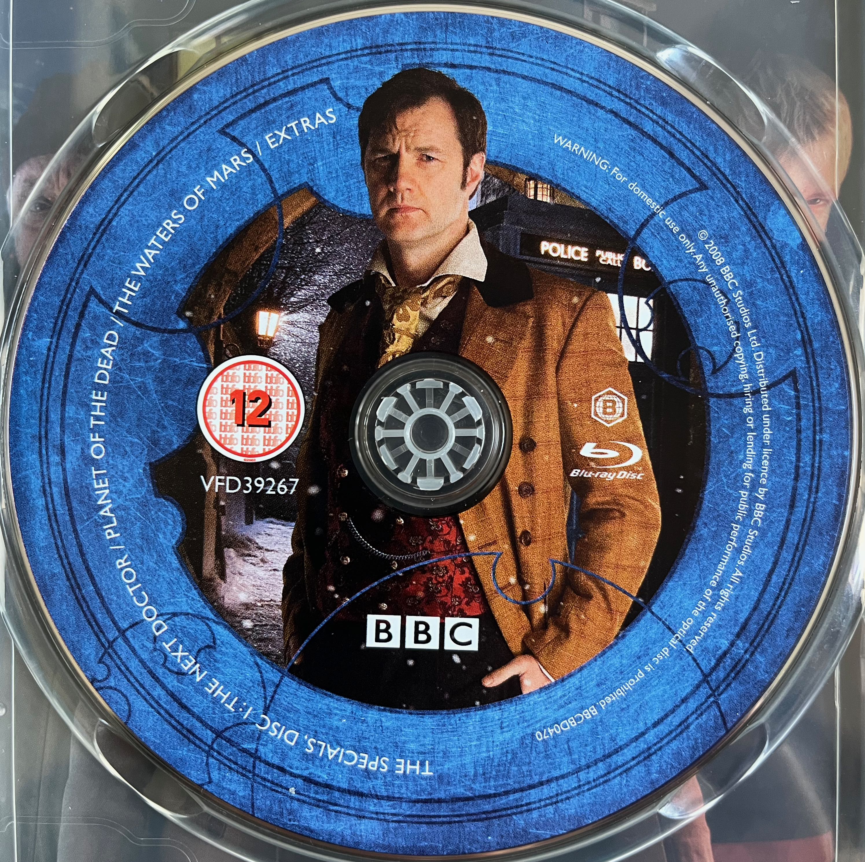 Disc 1 in the Doctor Who 2008 to 2010 Specials Blu-ray set. It features a photo of David Morrissey as Jackson Lake, who calls himself the Doctor, wearing a brown jacket and standing in front of the Tardis. A blue border around the edge of the disc contains the episode names and copyright information.