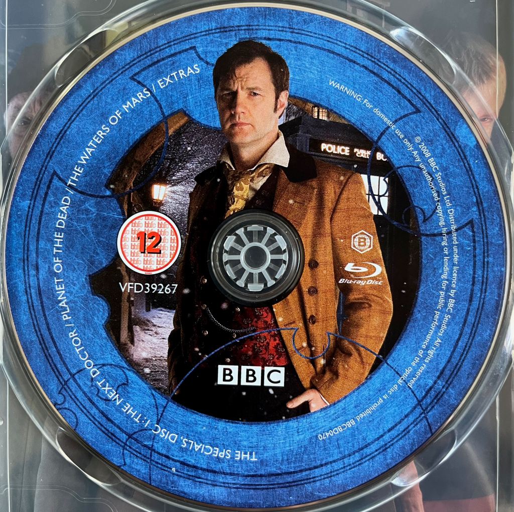 Disc 1 in the Doctor Who 2008 to 2010 Specials Blu-ray set. It features a photo of David Morrissey as Jackson Lake, who calls himself the Doctor, wearing a brown jacket and standing in front of the Tardis. A blue border around the edge of the disc contains the episode names and copyright information.